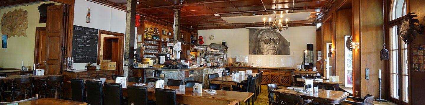 Ein gemütliches Restaurant mit Holztischen und -stühlen, einem Barbereich, einem großen Wandgemälde einer Sonnenbrillenträgerin an der Rückwand und warmer Beleuchtung durch Kronleuchter und Fenster.
